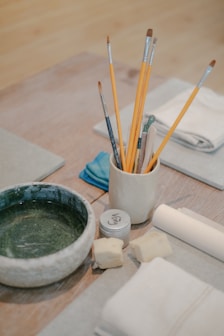 a wooden table topped with a bowl of paint and brushes