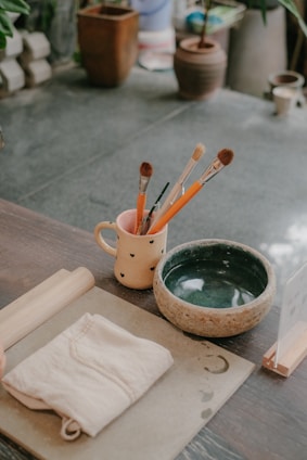An assortment of painting and crafting tools is arranged on a wooden table. A cream-colored mug with a heart design holds several paintbrushes and pencils. Next to the mug is a greenish bowl, and a folded cloth lies beside it. There are also a cylindrical wooden rolling pin and a flat board with water stains and a crescent shape. In the background, various potted plants can be seen on a stone floor.