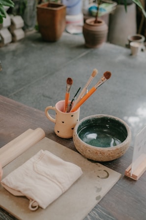 An assortment of painting and crafting tools is arranged on a wooden table. A cream-colored mug with a heart design holds several paintbrushes and pencils. Next to the mug is a greenish bowl, and a folded cloth lies beside it. There are also a cylindrical wooden rolling pin and a flat board with water stains and a crescent shape. In the background, various potted plants can be seen on a stone floor.