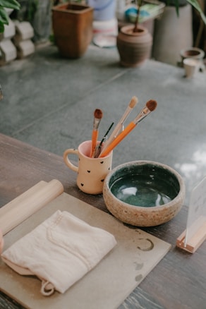 An assortment of painting and crafting tools is arranged on a wooden table. A cream-colored mug with a heart design holds several paintbrushes and pencils. Next to the mug is a greenish bowl, and a folded cloth lies beside it. There are also a cylindrical wooden rolling pin and a flat board with water stains and a crescent shape. In the background, various potted plants can be seen on a stone floor.