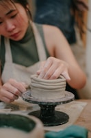 A person is working on a piece of pottery using the coil method, skillfully shaping the clay with their hands on a potter's wheel. The person is wearing an apron, indicating involvement in a hands-on craft activity.