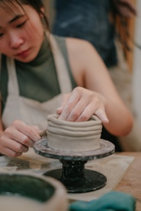 A person is working on a piece of pottery using the coil method, skillfully shaping the clay with their hands on a potter's wheel. The person is wearing an apron, indicating involvement in a hands-on craft activity.