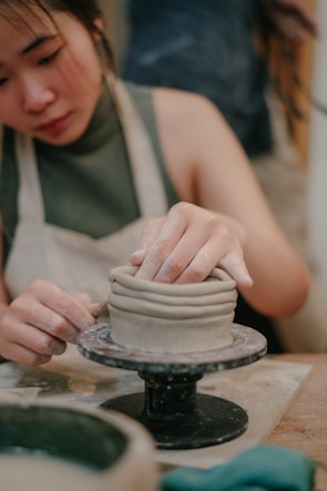 A person is working on a piece of pottery using the coil method, skillfully shaping the clay with their hands on a potter's wheel. The person is wearing an apron, indicating involvement in a hands-on craft activity.