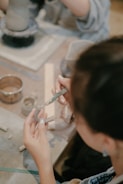 Hands are crafting with clay on a table, surrounded by various pottery tools and partially formed clay pieces. The focus is on the delicate shaping of clay with both hands and the use of a sculpting tool.