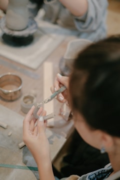 Hands are crafting with clay on a table, surrounded by various pottery tools and partially formed clay pieces. The focus is on the delicate shaping of clay with both hands and the use of a sculpting tool.