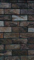 A close-up view of a brick wall composed of rectangular stones with a rough texture. The bricks vary in color, featuring shades of brown, gray, and beige. The wall is neatly arranged in horizontal rows, with each row offset from the one below it.