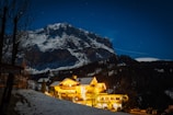 A snow-capped mountain looms in the background under a clear night sky filled with stars. In the foreground, a warmly lit hotel glows against the dark landscape, surrounded by trees and patches of snow. The scene is peaceful with contrasts of warm lights and cool mountain blues.