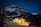A snow-capped mountain looms in the background under a clear night sky filled with stars. In the foreground, a warmly lit hotel glows against the dark landscape, surrounded by trees and patches of snow. The scene is peaceful with contrasts of warm lights and cool mountain blues.