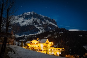 A snow-capped mountain looms in the background under a clear night sky filled with stars. In the foreground, a warmly lit hotel glows against the dark landscape, surrounded by trees and patches of snow. The scene is peaceful with contrasts of warm lights and cool mountain blues.