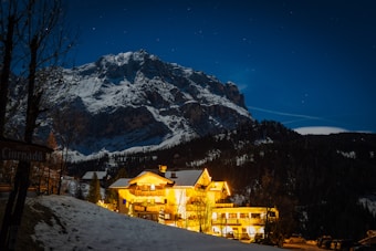 A snow-capped mountain looms in the background under a clear night sky filled with stars. In the foreground, a warmly lit hotel glows against the dark landscape, surrounded by trees and patches of snow. The scene is peaceful with contrasts of warm lights and cool mountain blues.