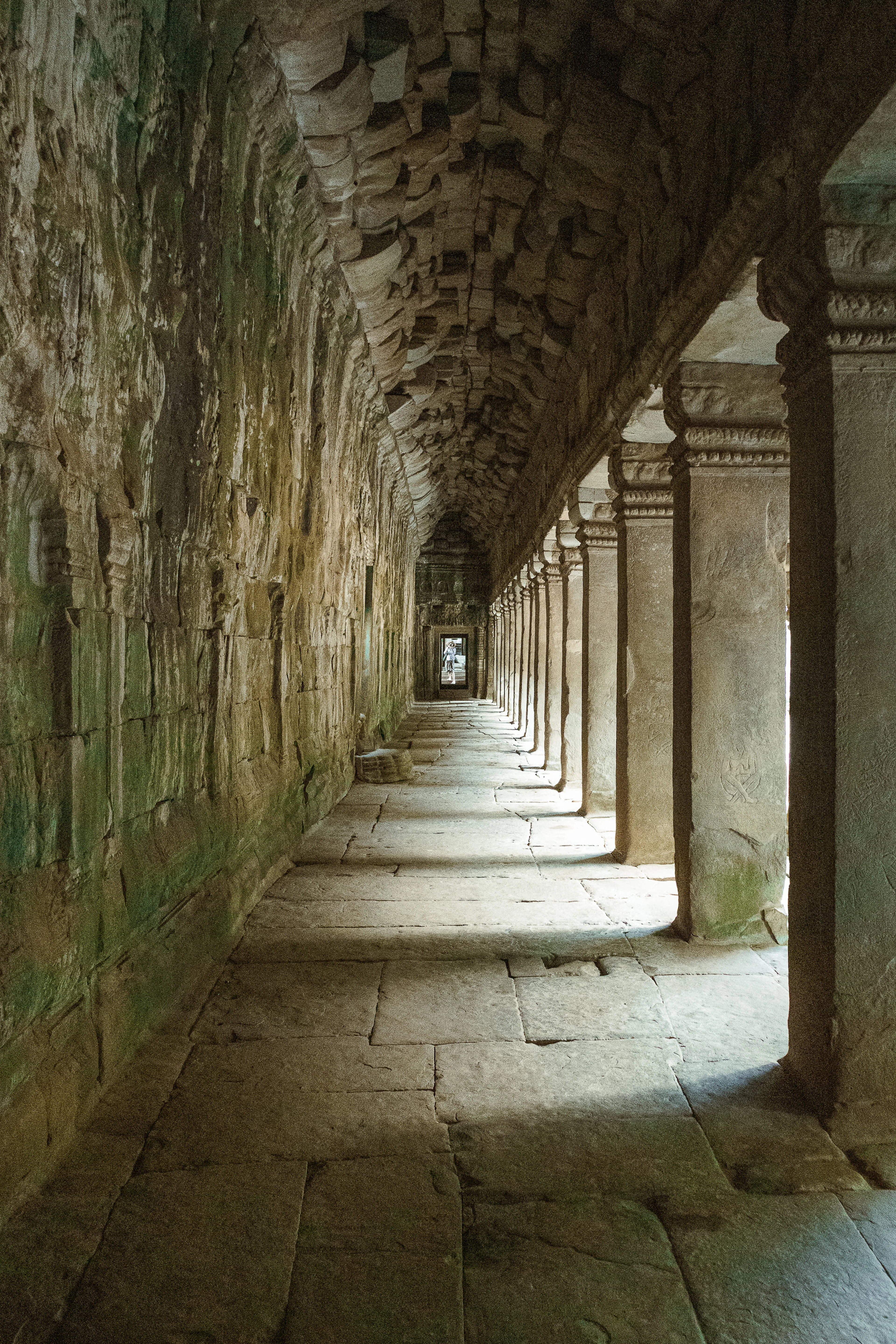 A long covered gallery inside an Angkor Thom temple, its corbelled stone vault running away into shadow above a row of carved pillars and a moss-stained wall