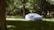 A car covered with a white protective tarp is parked on a grassy area surrounded by dense, green trees and vegetation. The scene is partially shaded by the foliage, creating a peaceful and secluded atmosphere.