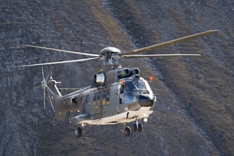 A military helicopter is flying over a mountainous landscape. The helicopter is large and prominently detailed with various markings and accessories.