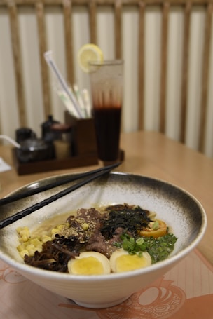 A bowl of ramen is presented with various toppings including boiled eggs, corn, seaweed, and slices of meat. Chopsticks are resting on the bowl, and a tall drink garnished with a lemon slice is in the background. The setting appears to be a restaurant table, with condiment containers visible.