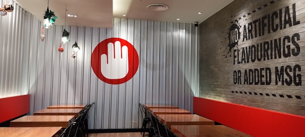 A restaurant interior featuring a series of wooden tables aligned against metallic corrugated walls, with black chairs neatly placed. A large red and white hand symbol decorates the wall on one side. On the adjacent wall, a message reads 'No artificial flavourings or added MSG' in bold black letters. A few small plants hang from the ceiling as decoration.