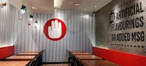 A restaurant interior featuring a series of wooden tables aligned against metallic corrugated walls, with black chairs neatly placed. A large red and white hand symbol decorates the wall on one side. On the adjacent wall, a message reads 'No artificial flavourings or added MSG' in bold black letters. A few small plants hang from the ceiling as decoration.