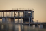 An unfinished multi-story building under construction with scaffolding and safety netting surrounding the structure. The scene appears to be taken during sunset or sunrise, casting a warm, golden hue across the site. Metal pipes, concrete pillars, and temporary platforms are visible, along with partially completed walls and floors.