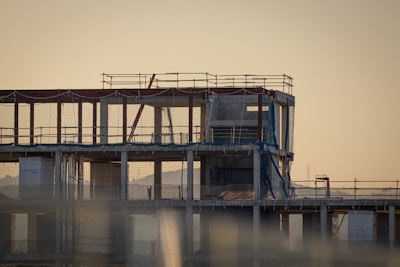 An unfinished multi-story building under construction with scaffolding and safety netting surrounding the structure. The scene appears to be taken during sunset or sunrise, casting a warm, golden hue across the site. Metal pipes, concrete pillars, and temporary platforms are visible, along with partially completed walls and floors.