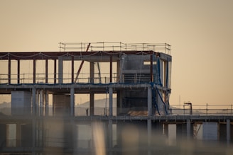 An unfinished multi-story building under construction with scaffolding and safety netting surrounding the structure. The scene appears to be taken during sunset or sunrise, casting a warm, golden hue across the site. Metal pipes, concrete pillars, and temporary platforms are visible, along with partially completed walls and floors.