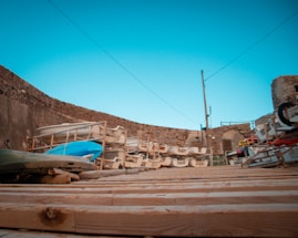 A stack of boats and kayaks stored on wooden racks in an outdoor area surrounded by stone walls. The foreground features a wooden deck, and in the background, there is a clear blue sky.