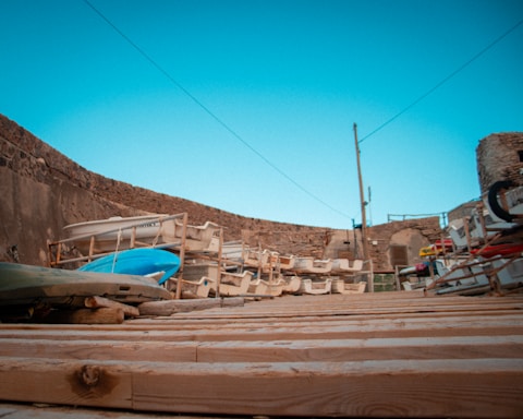 A stack of boats and kayaks stored on wooden racks in an outdoor area surrounded by stone walls. The foreground features a wooden deck, and in the background, there is a clear blue sky.