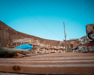 A stack of boats and kayaks stored on wooden racks in an outdoor area surrounded by stone walls. The foreground features a wooden deck, and in the background, there is a clear blue sky.