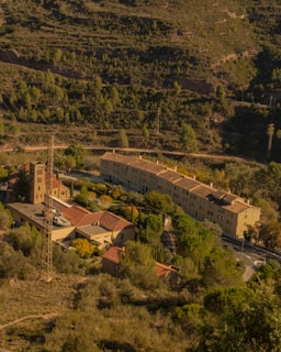 An aerial view of a small village nestled in a hilly, forested area. The image features several buildings, including a church with a bell tower and residential structures with red-tiled roofs. The landscape is lush with greenery, and a narrow road winds around the village.