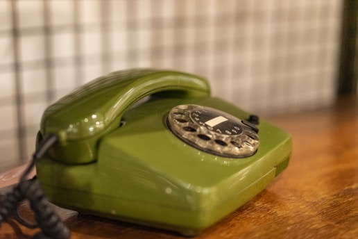 A vintage green rotary dial telephone is placed on a wooden surface with a blurred, patterned background. The phone features a coiled cord and a classic dial with numbers visible.