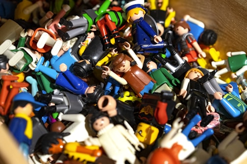 A collection of colorful plastic toy figures scattered in disarray. Various figures are dressed in different outfits, representing a diverse array of themes and professions. The toys are made of bright plastic, with some figures facing upwards and others sideways or upside down. The scene appears playful and chaotic.