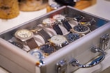 A collection of wristwatches arranged neatly inside a transparent display case. The watches have various designs and face colors, with their metallic and leather straps visible. The case is set on a light surface, and there are blurred objects in the background, suggesting a setting of display or storage.