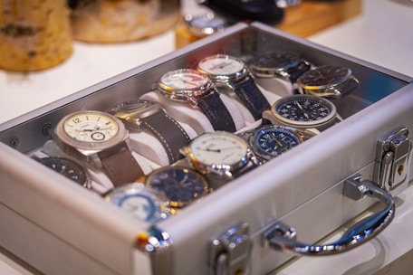 A collection of wristwatches arranged neatly inside a transparent display case. The watches have various designs and face colors, with their metallic and leather straps visible. The case is set on a light surface, and there are blurred objects in the background, suggesting a setting of display or storage.