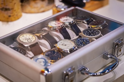 A collection of wristwatches arranged neatly inside a transparent display case. The watches have various designs and face colors, with their metallic and leather straps visible. The case is set on a light surface, and there are blurred objects in the background, suggesting a setting of display or storage.