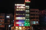 A multi-story hotel building is lit up at night with various colored lights. The facade displays the hotel name 'SDM Vedanta' and the sign '24x7 Restaurant' at the top. The building features a modern design with glass windows and a combination of warm and cool lighting. Adjacent structures are partially visible.