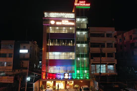 A multi-story hotel building is lit up at night with various colored lights. The facade displays the hotel name 'SDM Vedanta' and the sign '24x7 Restaurant' at the top. The building features a modern design with glass windows and a combination of warm and cool lighting. Adjacent structures are partially visible.