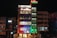 A multi-story hotel building is lit up at night with various colored lights. The facade displays the hotel name 'SDM Vedanta' and the sign '24x7 Restaurant' at the top. The building features a modern design with glass windows and a combination of warm and cool lighting. Adjacent structures are partially visible.