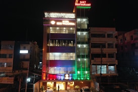 A multi-story hotel building is lit up at night with various colored lights. The facade displays the hotel name 'SDM Vedanta' and the sign '24x7 Restaurant' at the top. The building features a modern design with glass windows and a combination of warm and cool lighting. Adjacent structures are partially visible.