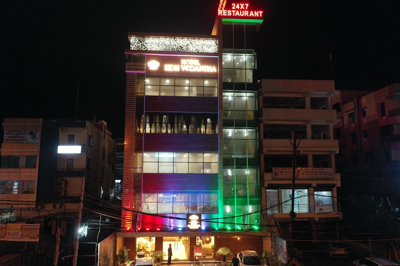A multi-story hotel building is lit up at night with various colored lights. The facade displays the hotel name 'SDM Vedanta' and the sign '24x7 Restaurant' at the top. The building features a modern design with glass windows and a combination of warm and cool lighting. Adjacent structures are partially visible.