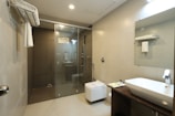 A modern bathroom features a glass-enclosed shower area with a mounted showerhead. A white ceramic sink rests on a dark wooden counter, alongside a mirror and toiletries. Towels hang on a rack above the commode, and the space is illuminated by ceiling lights.