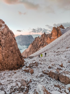 A rugged mountainous landscape with snow-covered rocky terrain. Several people are hiking in the distance, dressed in outdoor gear. The sky is partially cloudy, with the sun low on the horizon, casting a warm glow on the peaks.