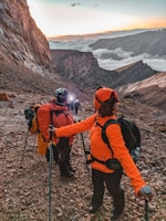 A group of hikers in colorful jackets ascend a rocky mountain trail. The sky is illuminated by a sunrise or sunset, with clouds nestled in the valley below. Participants carry hiking poles and backpacks, indicating a challenging expedition.