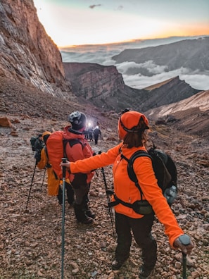 A group of hikers in colorful jackets ascend a rocky mountain trail. The sky is illuminated by a sunrise or sunset, with clouds nestled in the valley below. Participants carry hiking poles and backpacks, indicating a challenging expedition.