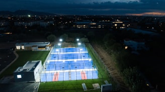 Aerial view of a brightly lit padel court complex at night, surrounded by greenery and nearby buildings. The courts are illuminated, creating a vivid contrast against the evening sky. In the distance, city lights and a mountain range are visible beneath a cloudy sky.