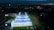 Aerial view of a brightly lit padel court complex at night, surrounded by greenery and nearby buildings. The courts are illuminated, creating a vivid contrast against the evening sky. In the distance, city lights and a mountain range are visible beneath a cloudy sky.