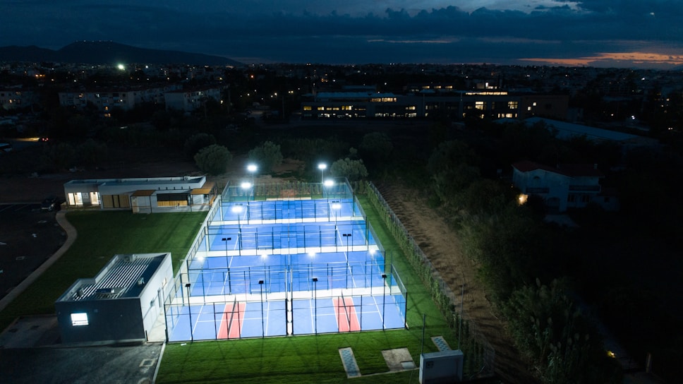 Aerial view of a brightly lit padel court complex at night, surrounded by greenery and nearby buildings. The courts are illuminated, creating a vivid contrast against the evening sky. In the distance, city lights and a mountain range are visible beneath a cloudy sky.