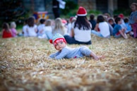 A young child lying on straw-covered ground wearing a festive hat with 'Santa's Helper' written on it. The background is filled with a group of people, mostly children, sitting together, some wearing Santa hats, suggesting a holiday gathering.