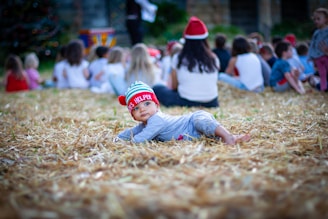 A young child lying on straw-covered ground wearing a festive hat with 'Santa's Helper' written on it. The background is filled with a group of people, mostly children, sitting together, some wearing Santa hats, suggesting a holiday gathering.