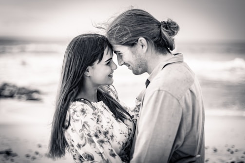 A black and white photo of a couple standing close together on a beach. They are smiling at each other, exuding warmth and affection. The background features a serene ocean and a cloudy sky, adding a gentle and calm atmosphere to the scene.