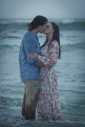 A couple shares a romantic kiss on the beach, with gentle waves in the background. The man is wearing a blue shirt and brown pants, while the woman is dressed in a long, floral-patterned dress. The setting is serene, with overcast skies.
