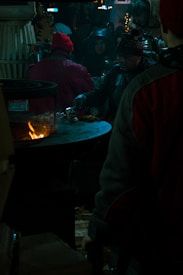 Several people gather around a large outdoor grill at night. Warm clothing suggests cold weather. A person is cooking food over an open flame while others wait nearby. Festive lights and decorations are visible, creating a cozy atmosphere.