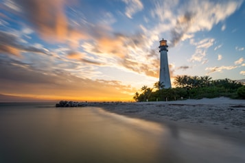 A tall lighthouse stands prominently against a backdrop of a vibrant sunset. The sky is filled with dynamic clouds reflecting hues of orange, pink, and blue. The lighthouse is surrounded by lush palm trees, and the view extends over a serene beach with gentle waves lapping at the shore.